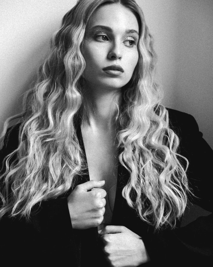 A woman with long, wavy hair poses thoughtfully against a neutral background. Her expression is serene, wearing a dark jacket, evoking a contemplative mood.